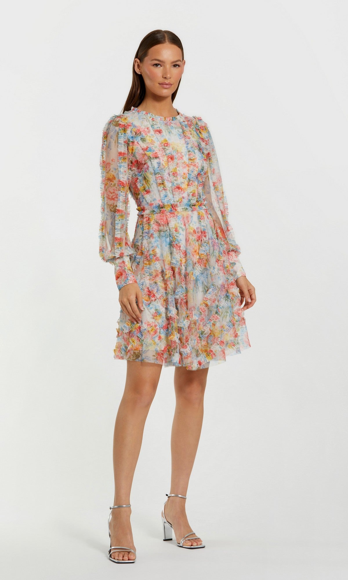 A woman stands against a light background wearing the Short Wedding Guest Dress: Mac Duggal 8304 in Blue Multi—a long-sleeve, floral mesh dress with a fitted waist—paired with silver high-heeled sandals.